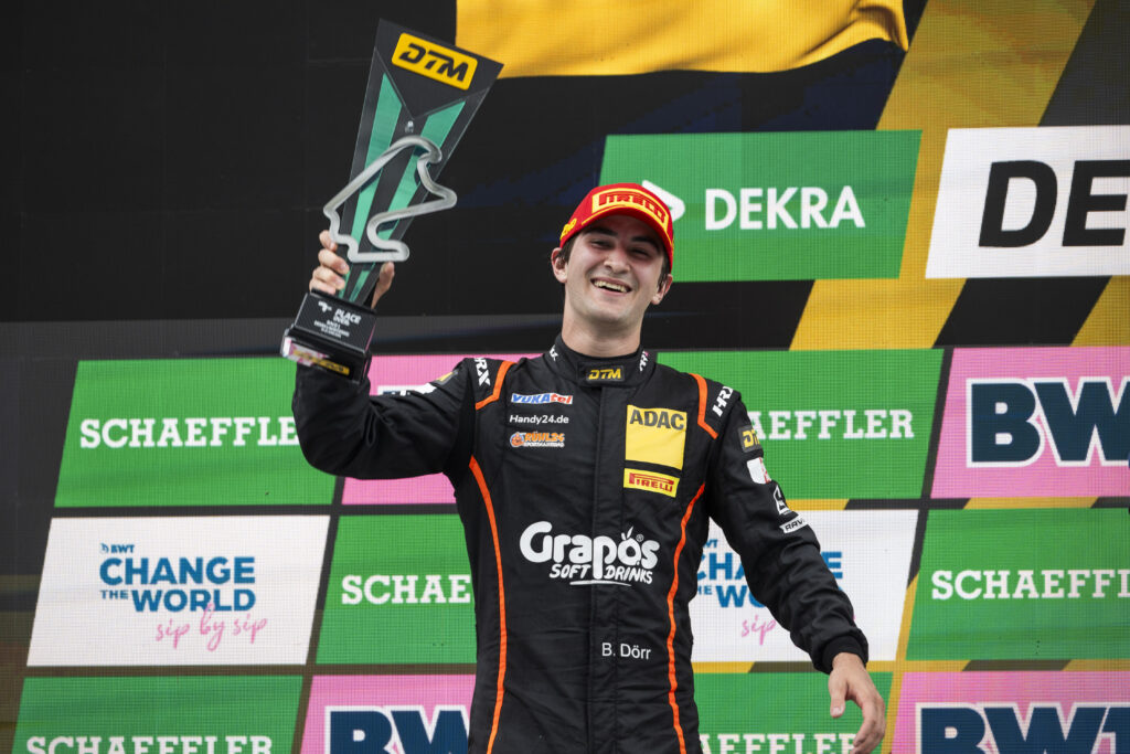 Rennfahrer Ben Dörr hält lächelnd eine DTM-Trophäe auf dem Podium hoch.