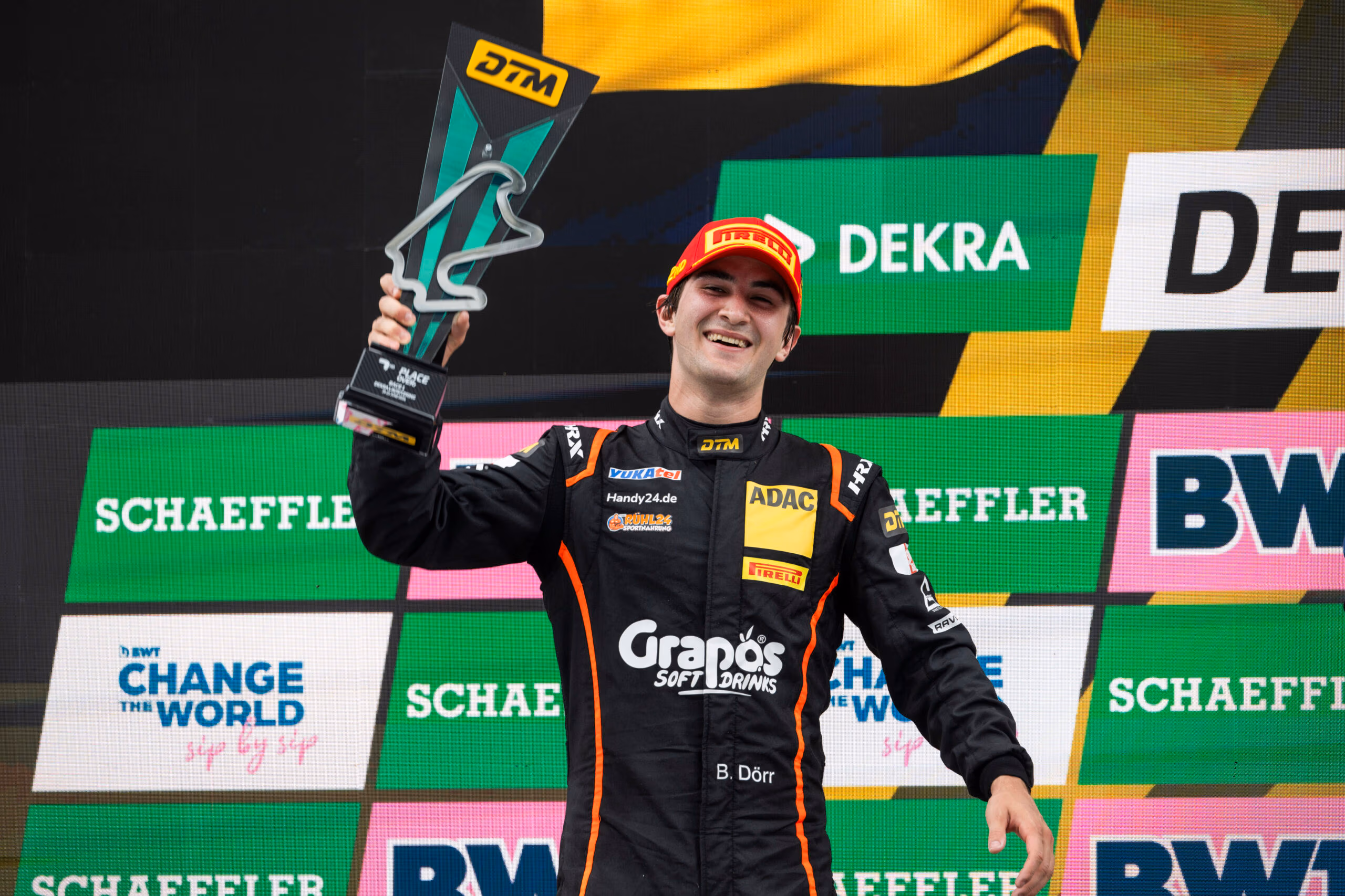 Rennfahrer Ben Dörr hält lächelnd einen DTM-Pokal vor einer Sponsorenwand hoch.